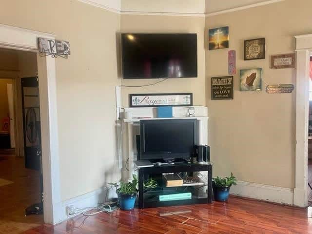 A living room corner with two mounted tvs, one above the other, on a beige wall with decorative signs and potted plants on a tv stand.