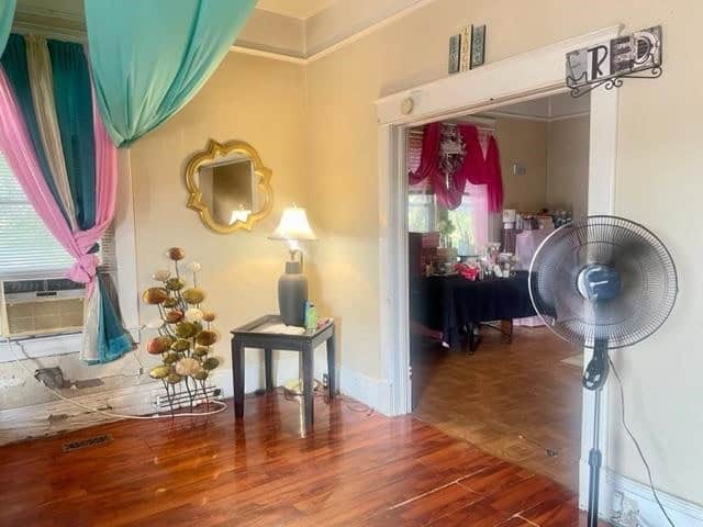 A vintage-inspired room featuring hardwood floors, an eclectic mix of colorful curtains, a decorative mirror on the wall, a standing fan, and a tree-shaped ornament beside a small table with a vase.