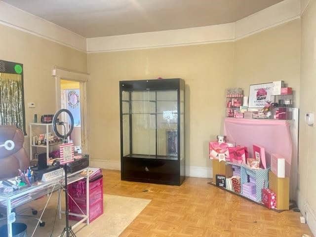 A room with beige walls and a parquet floor featuring a glass display cabinet, a table with cosmetics and a mirror, and shelves with various boxed products.