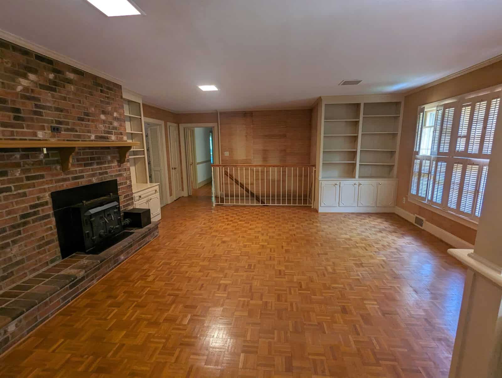 An empty room featuring a parquet floor, a brick fireplace, built-in bookshelves, and a safety gate.
