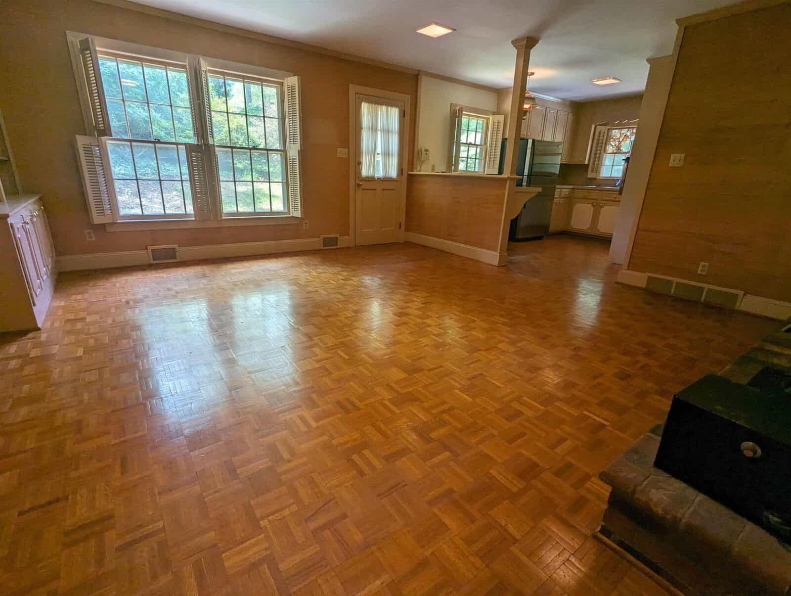 An empty room with parquet flooring, a radiant heating system, large windows, and an open connection to an adjacent kitchen area.