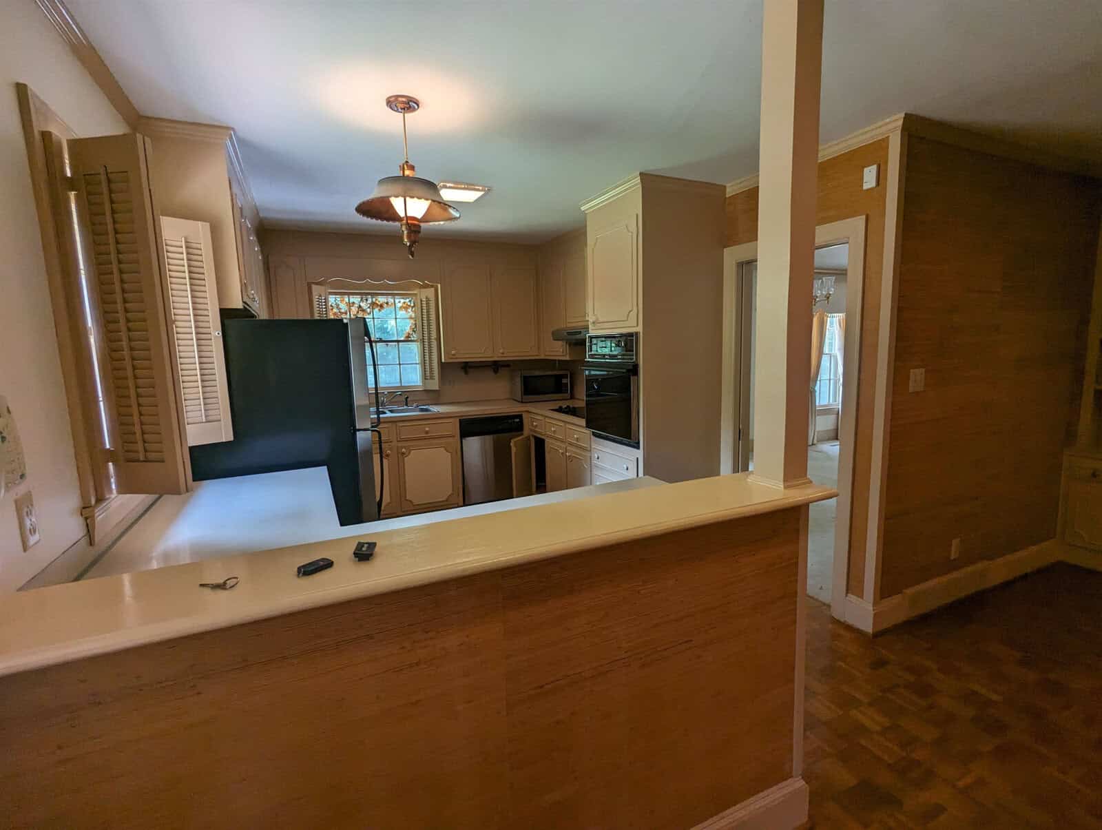 A domestic kitchen with wooden flooring, light beige cabinets, and standard appliances, viewed from an adjoining room.
