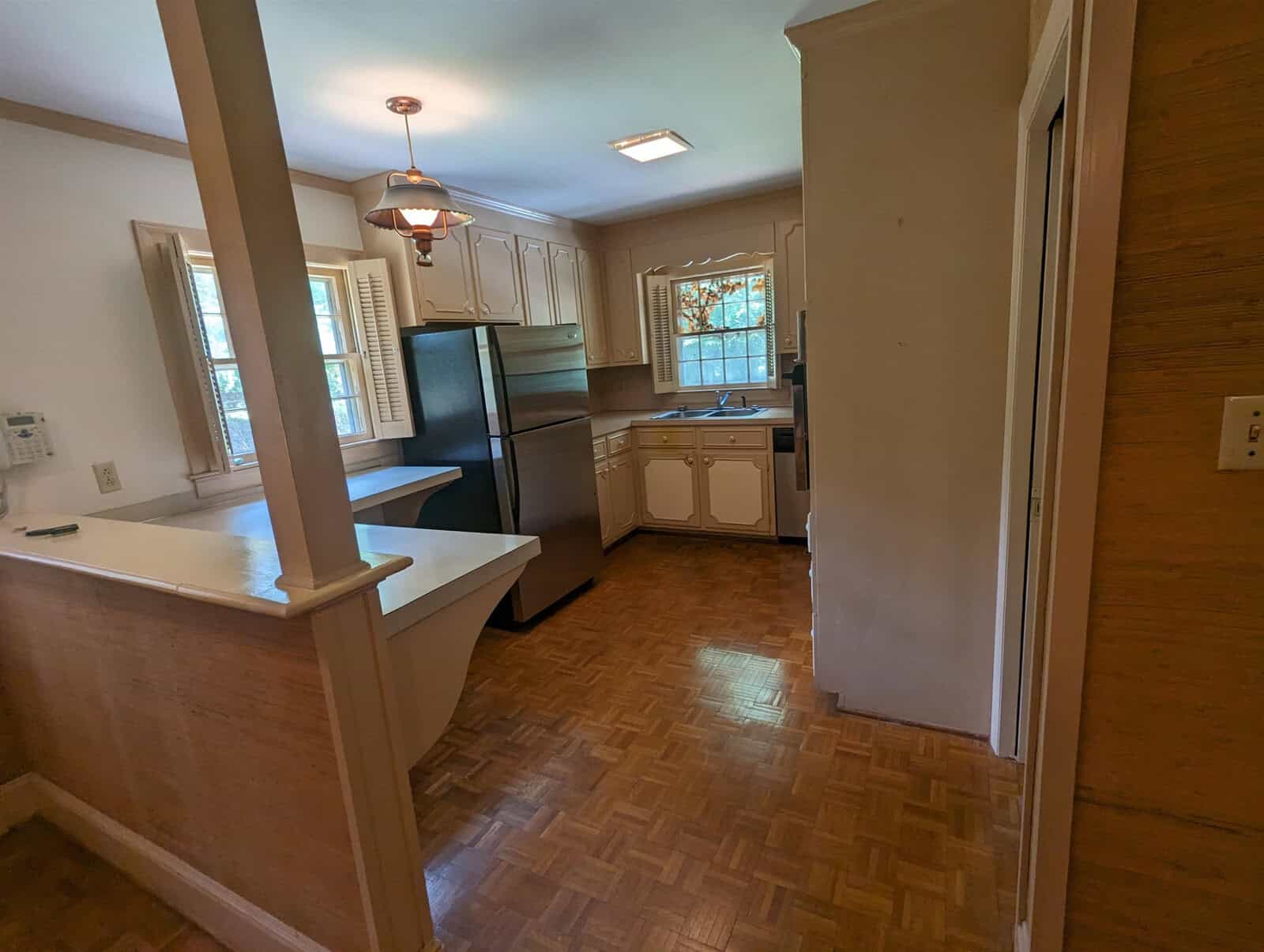 A view into a small, traditional kitchen with wood flooring, white appliances, and wooden cabinets, with natural light coming in from a window.
