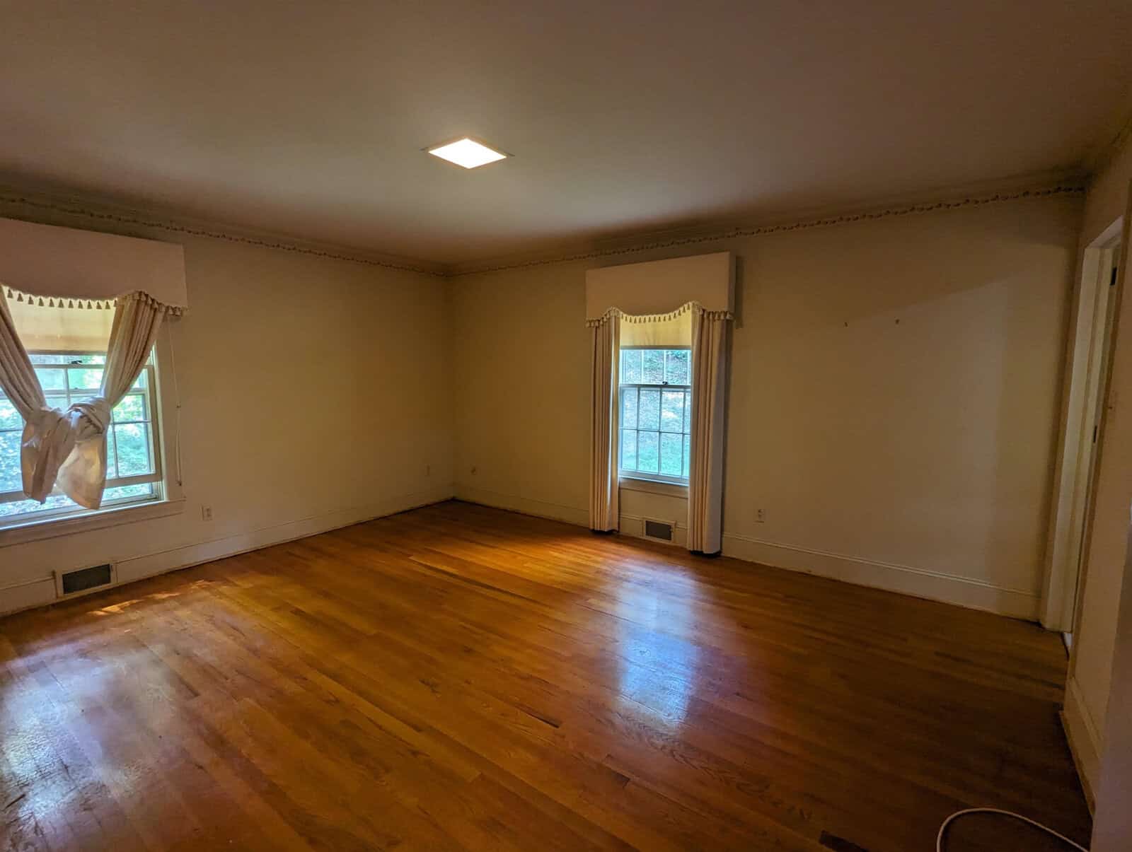 An empty room with hardwood floors, two windows with curtains, and a single overhead light fixture.