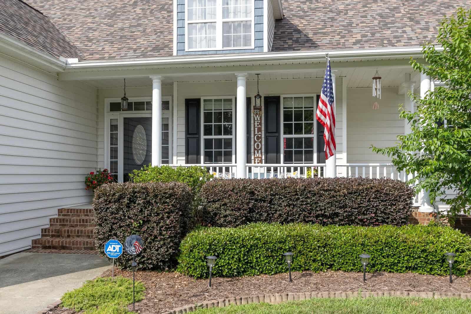Afront of a house showcasing a porch with columns, an american flag, a "welcome" sign, neatly trimmed hedges, and a home security sign on a sunny day.