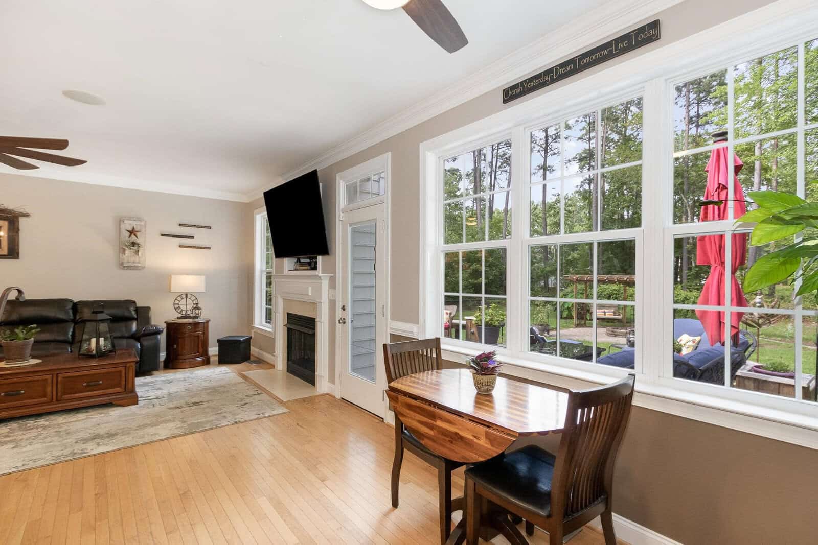 A well-lit, spacious living room with large windows, hardwood floors, and modern furnishings, including a dining table, chairs, and a wall-mounted television.