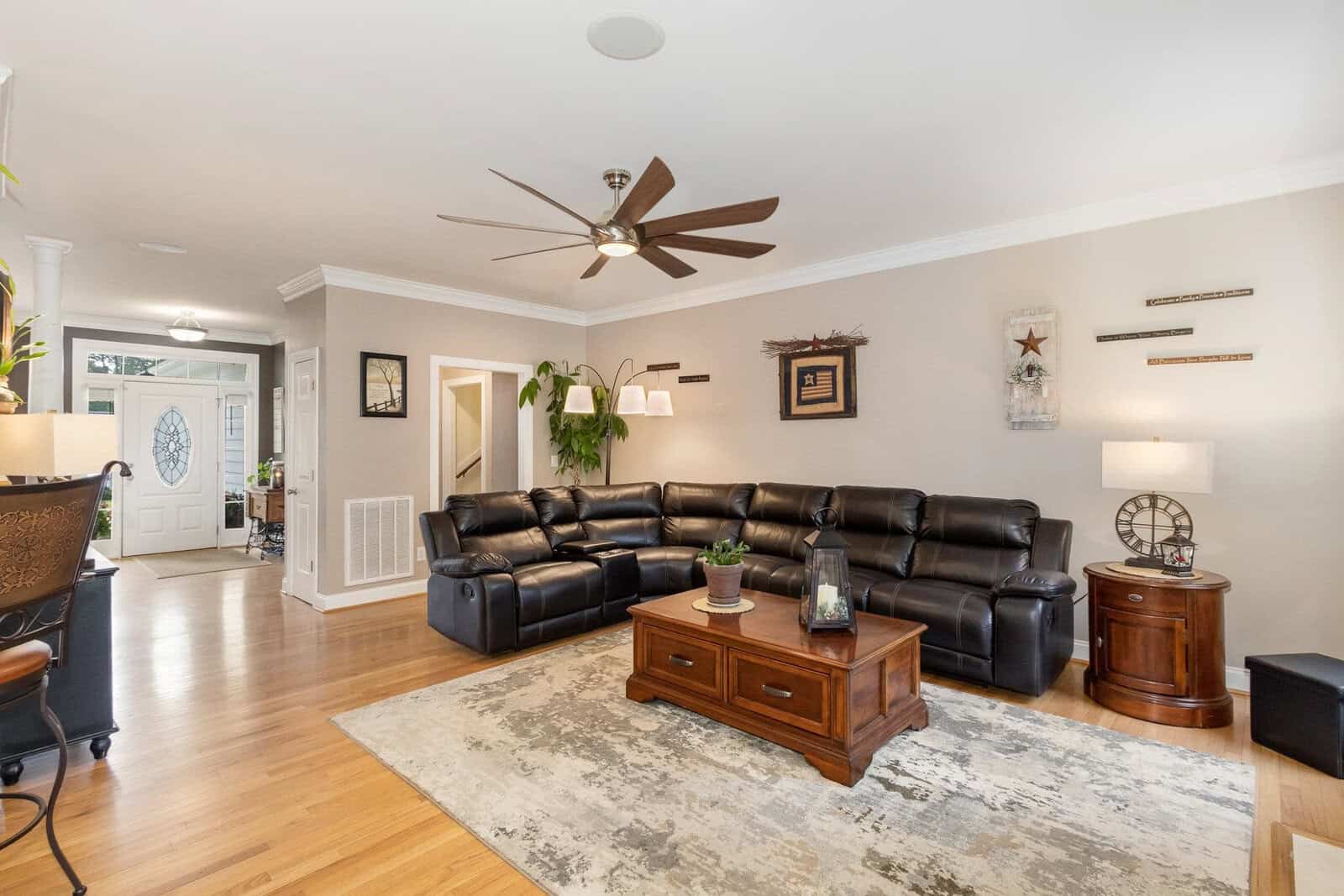 A well-lit living room featuring a large black leather sectional sofa, a wooden coffee table, a patterned area rug, and hardwood floors. decor includes a ceiling fan, wall-mounted shelves, and a variety of decorative items. the room opens into a hallway with a white door.