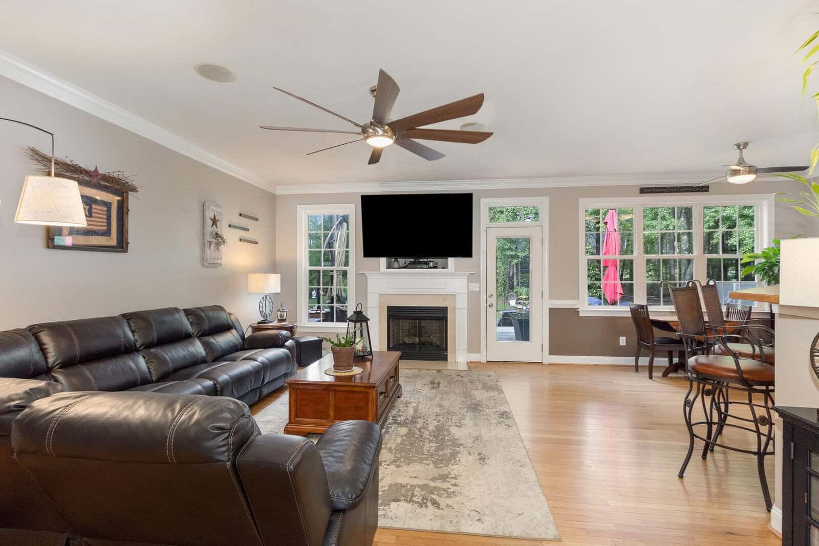 A well-lit living room with a dark leather sofa facing a mounted television above a fireplace, adjacent to a dining area with the door open to the outside.