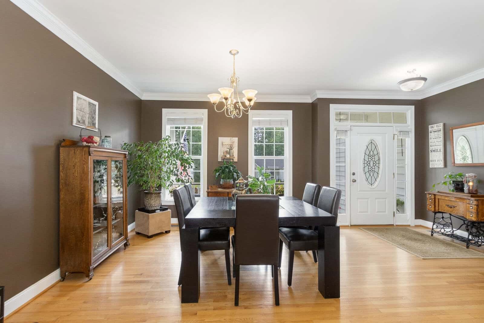 A spacious dining room featuring a dark wooden table with six chairs, hardwood floors, light grey walls, and a chandelier, with natural light streaming in through the windows and a glass-paneled door.
