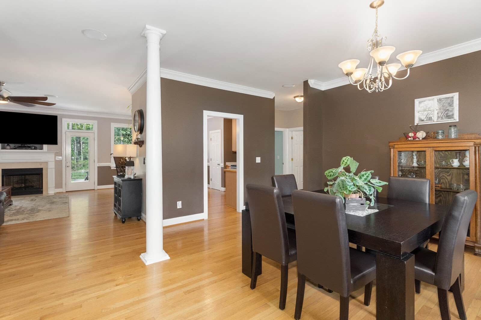 A spacious dining room featuring a dark wooden table with matching chairs, a fireplace, a chandelier, and white column details, with an adjoining room visible in the background.