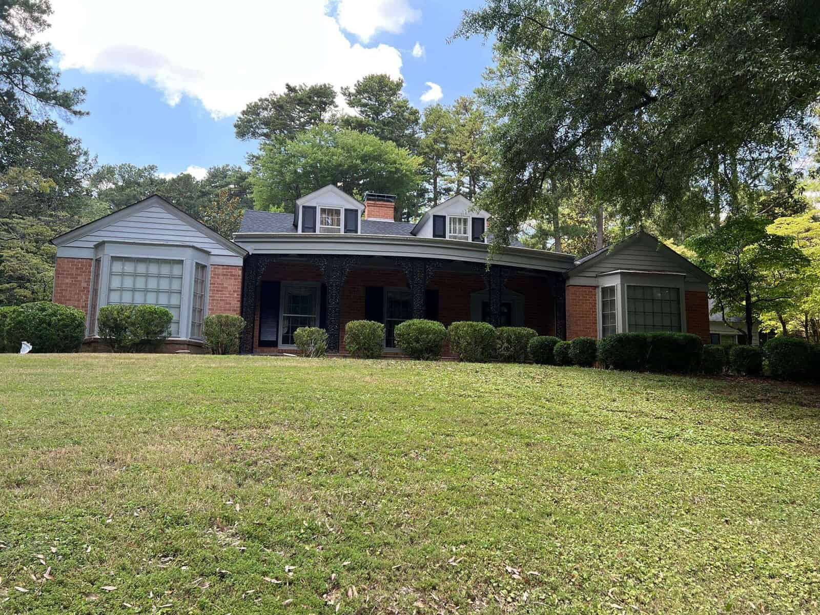 Single-story brick house with a gabled roof and dormer windows at 507 Granite Street, Henderson NC, flanked by two bay windows, set against a backdrop of trees and a clear sky,