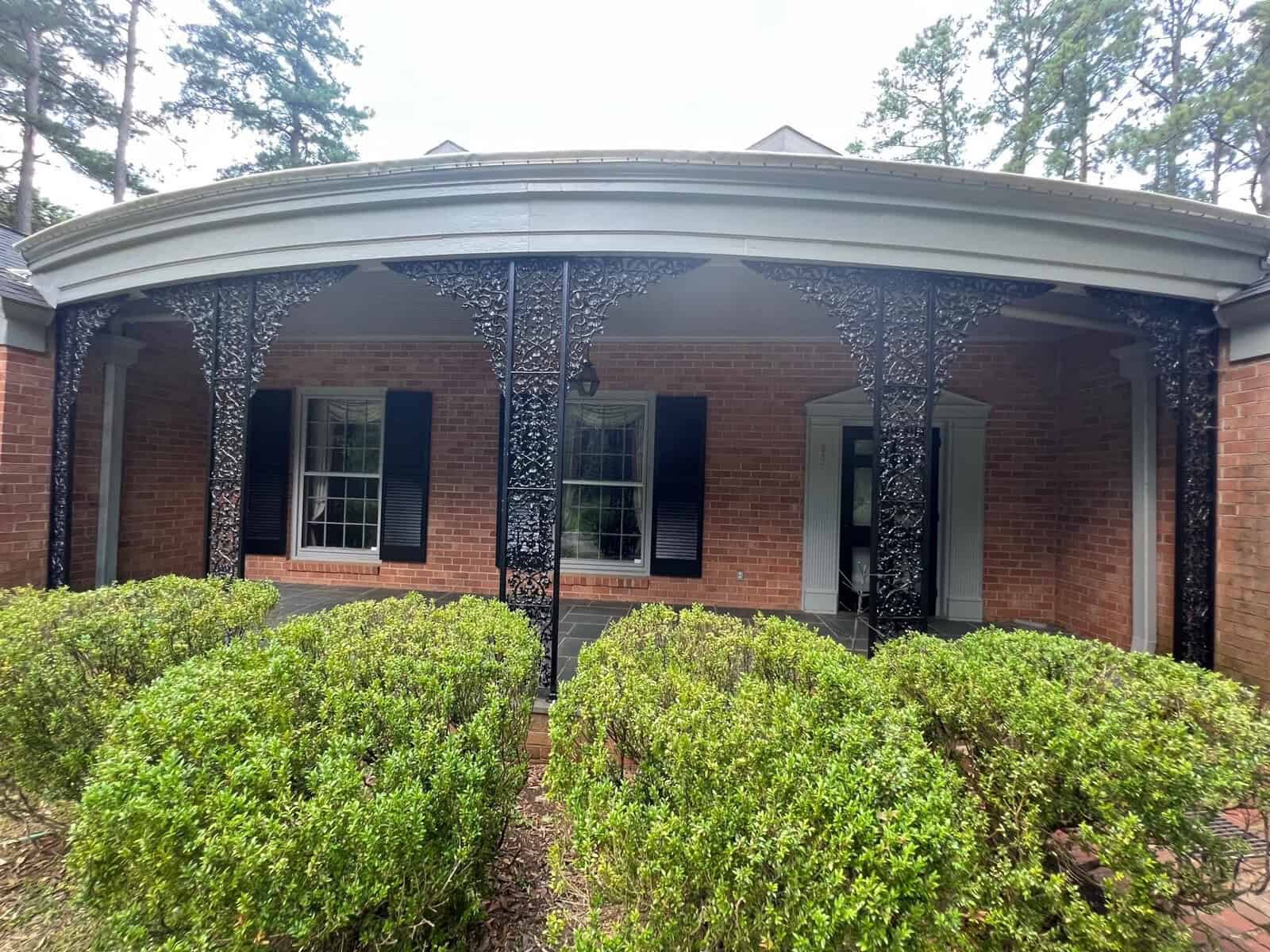 A red brick building with a curved porch supported by intricate metal columns, framed by green shrubbery in the foreground.