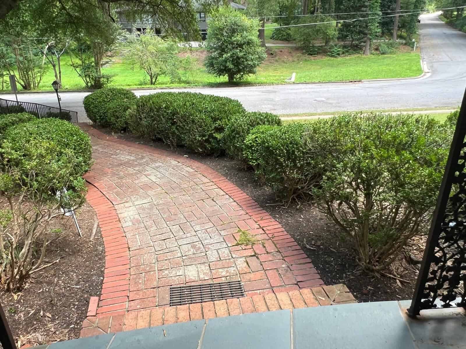A brick walkway leading down from a porch, flanked by manicured shrubs, with a street and grassy area visible in the background.