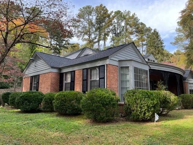 A single-story brick house with dark shutters, a gray shingled roof, and surrounded by green shrubbery, with trees displaying autumn foliage in the background.
