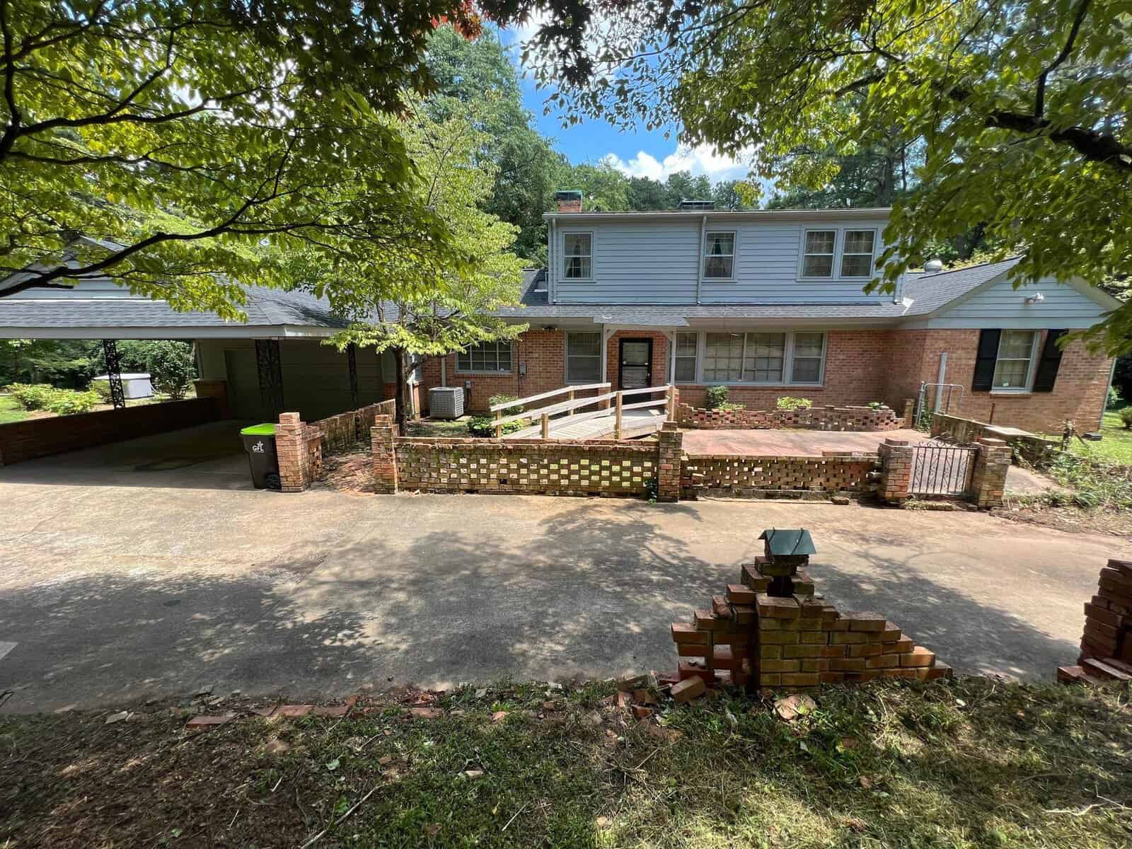 A brick house with a white roof and a wooden deck, viewed from an elevated angle on a sunny day, with surrounding greenery and a broad driveway leading to a carport.