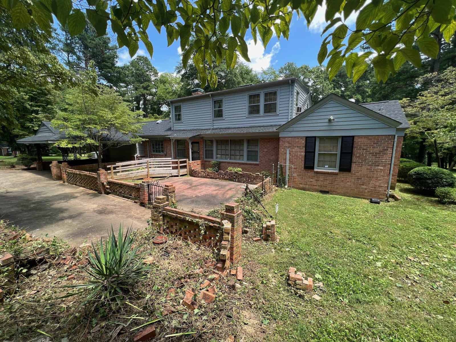 Two-story brick house with a wooden deck and a damaged front brick staircase, surrounded by greenery, under a clear sky.