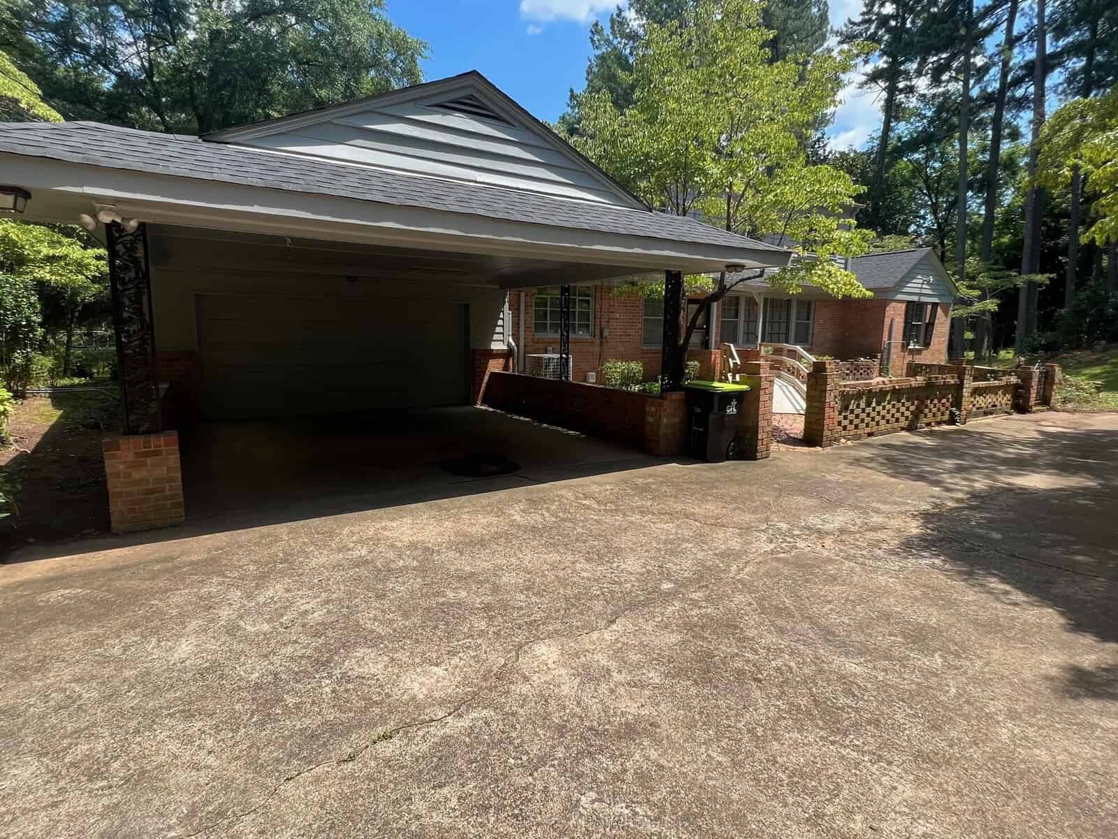A suburban brick house with an attached carport and a paved driveway, surrounded by trees on a sunny day.
