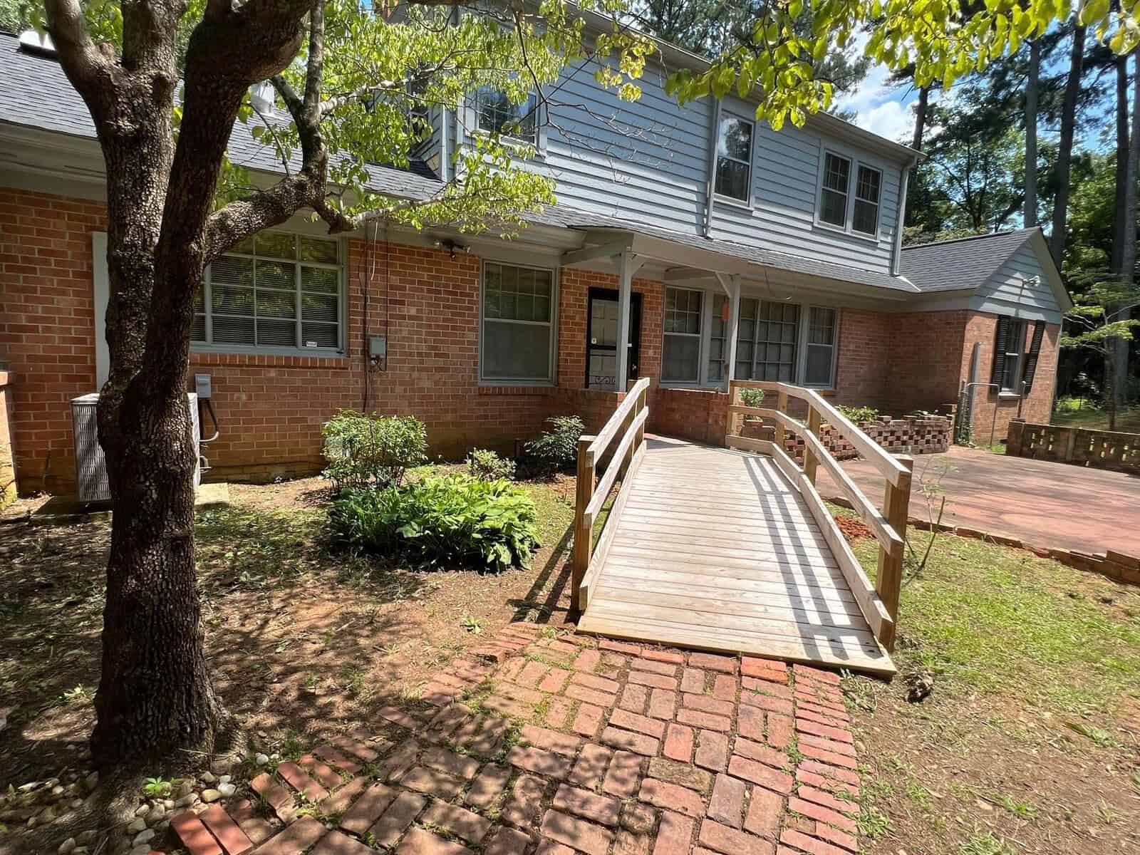 A brick and siding house with a new wooden ramp leading to the front door, surrounded by trees and landscaping on a sunny day.