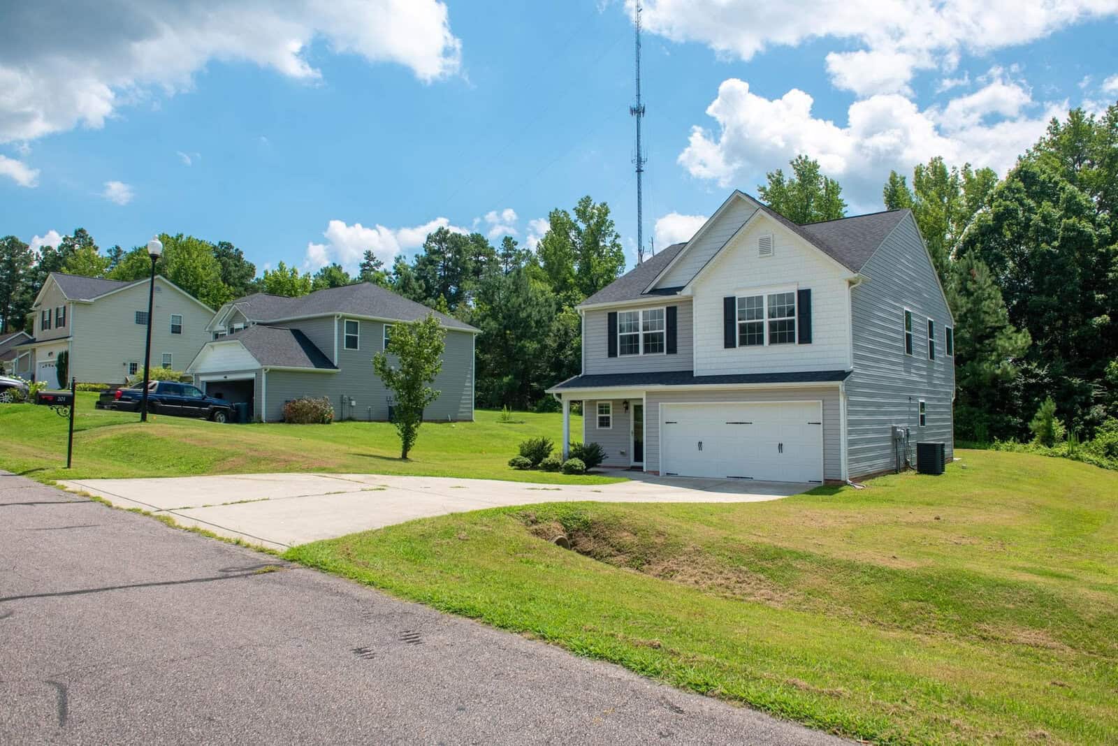 A suburban neighborhood with a two-story house featuring a garage, situated on a sloped lawn under a clear sky.