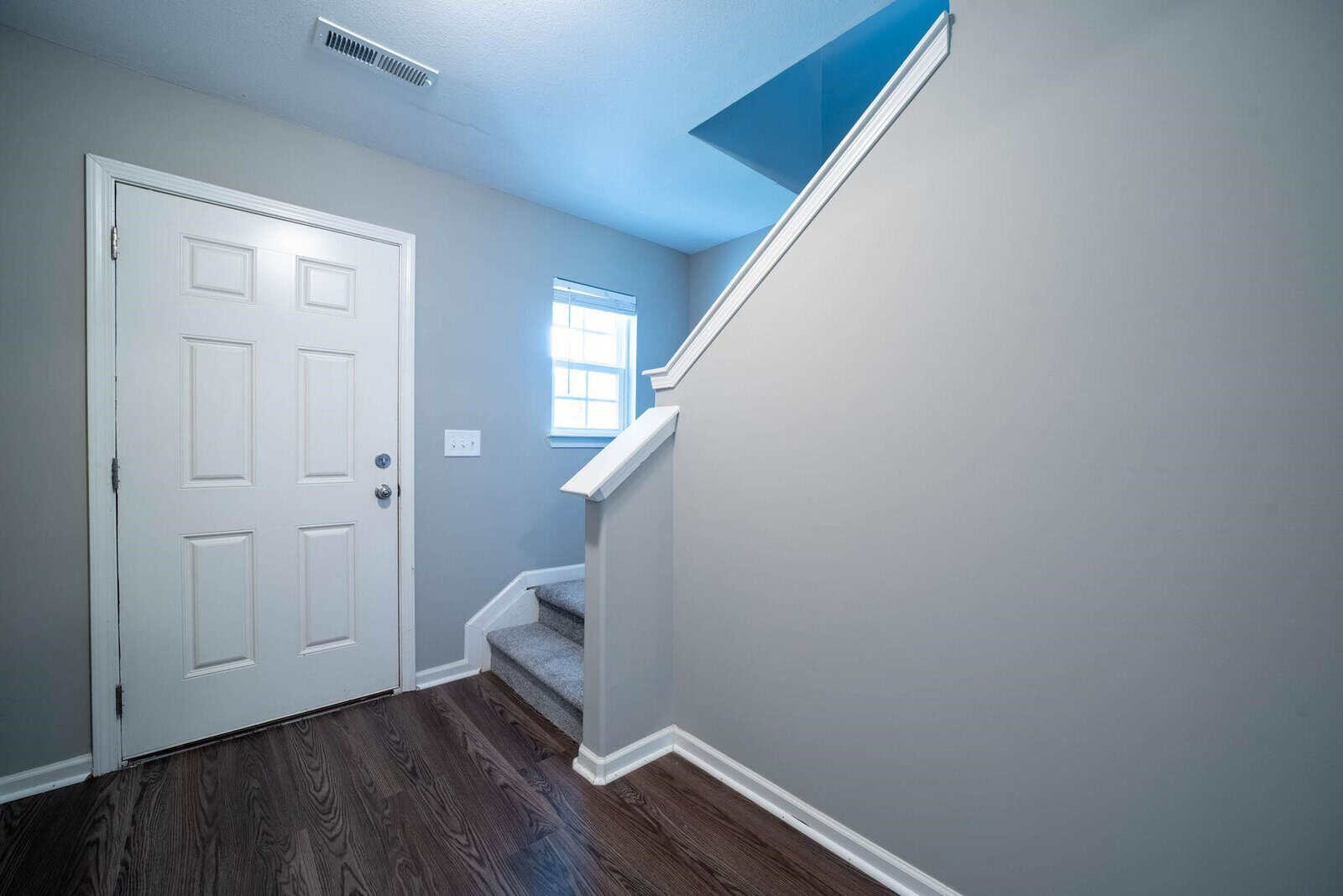 A clean and empty room with white walls, showing a closed white door on the left and a staircase with a white handrail leading up on the right, with natural light coming in through a small window.
