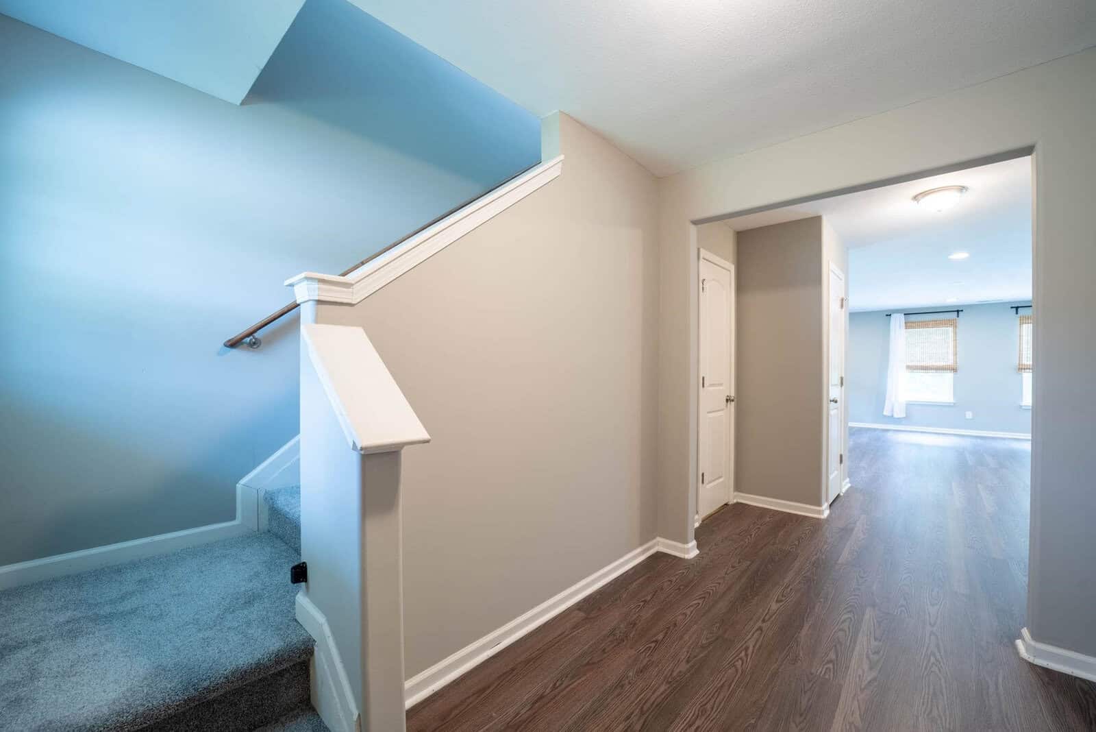 Interior of a modern home featuring a staircase with gray carpeting on the steps, white banister, and wooden flooring in the upper hallway, with light blue walls and white trim. a door is visible on the right, leading into another room.