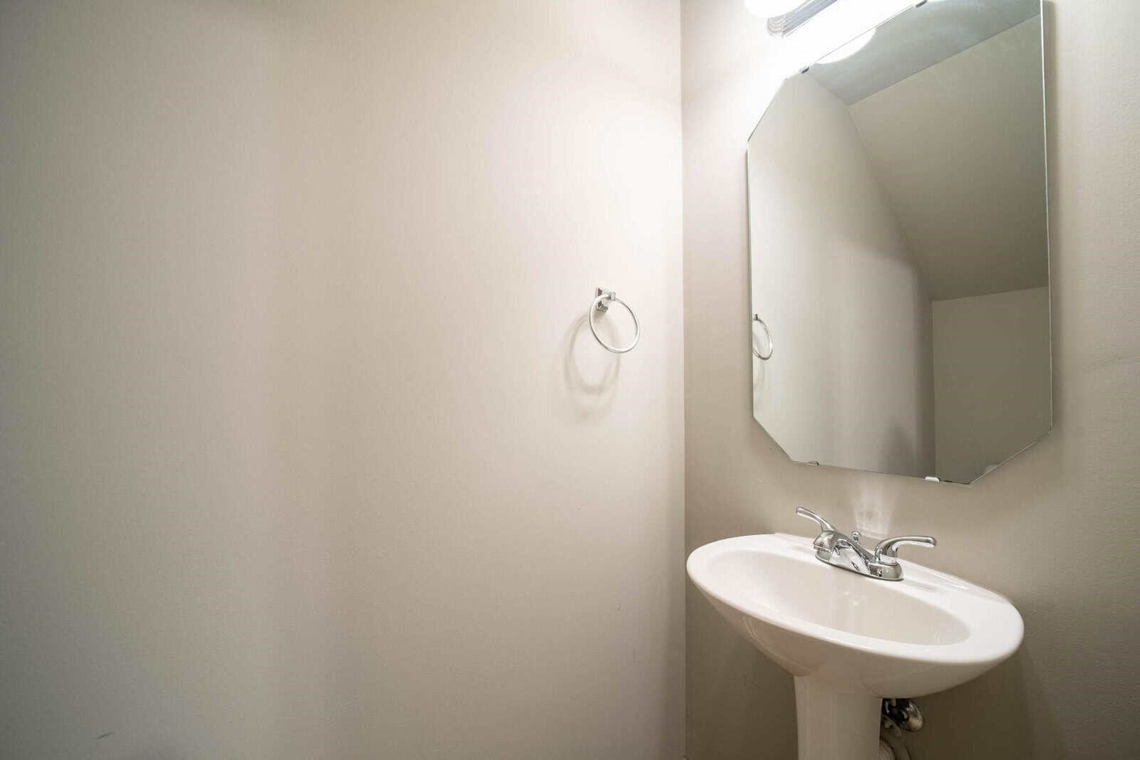 A corner of a bathroom featuring a white pedestal sink with a chrome faucet and a wall-mounted mirror above it, alongside a towel ring installed on the adjacent wall, all illuminated by a bright light source.