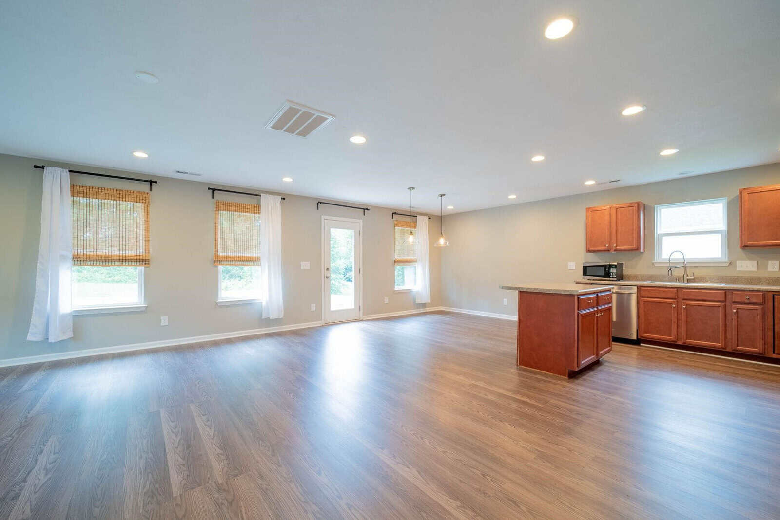 A spacious, empty room with wooden flooring, featuring a kitchen area with wooden cabinets and modern appliances, lit by natural light from windows with blinds and several recessed ceiling lights.