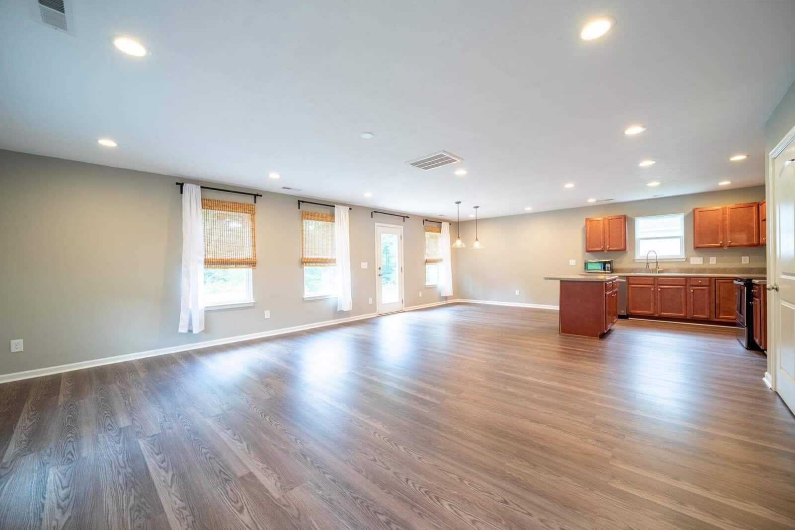 Spacious empty room with hardwood flooring, multiple windows with light curtains, and an adjoining kitchen area with wooden cabinets.