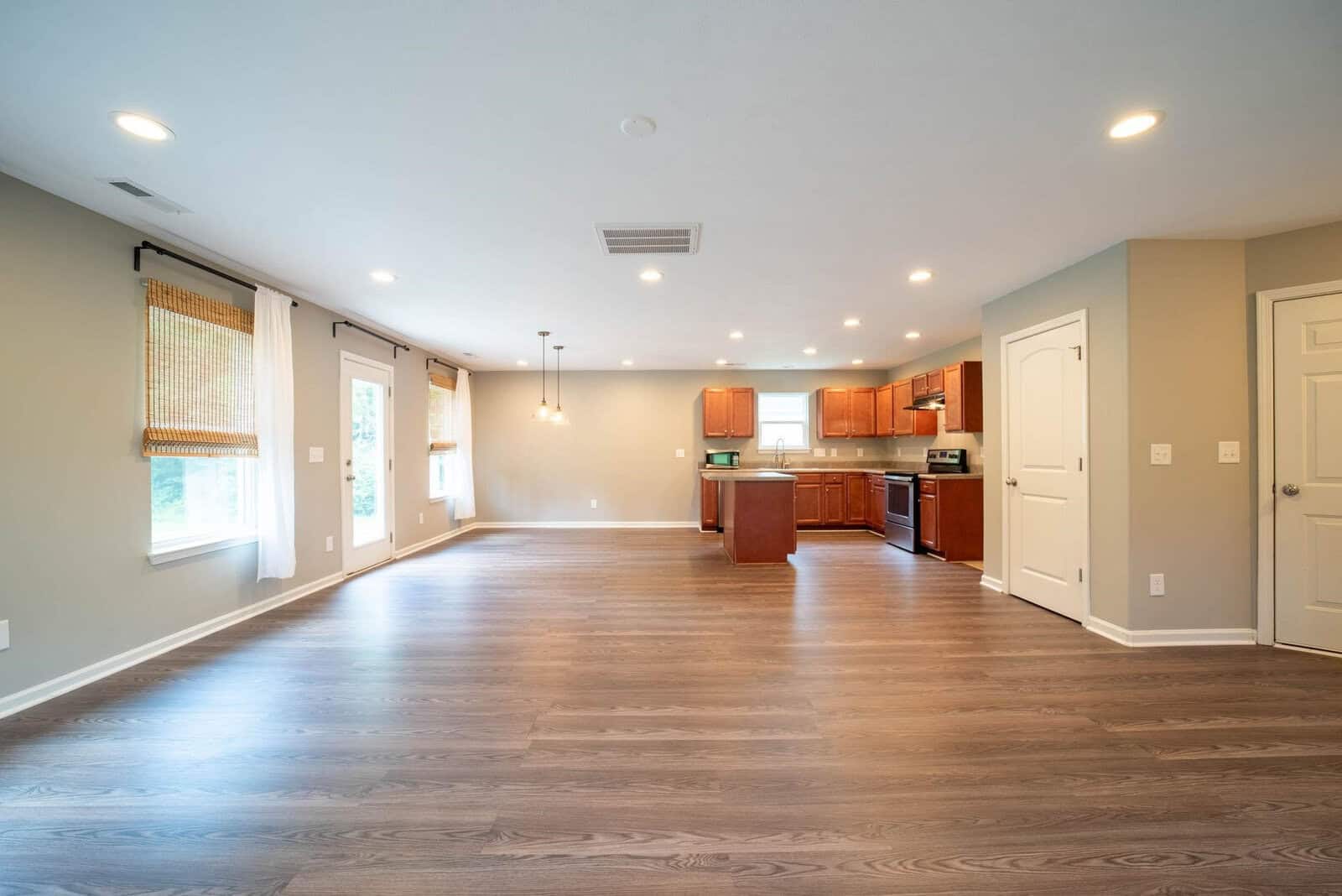 Open-plan living space with hardwood flooring, featuring a kitchen with wooden cabinets and stainless steel appliances, leading into a spacious room with ample natural light.