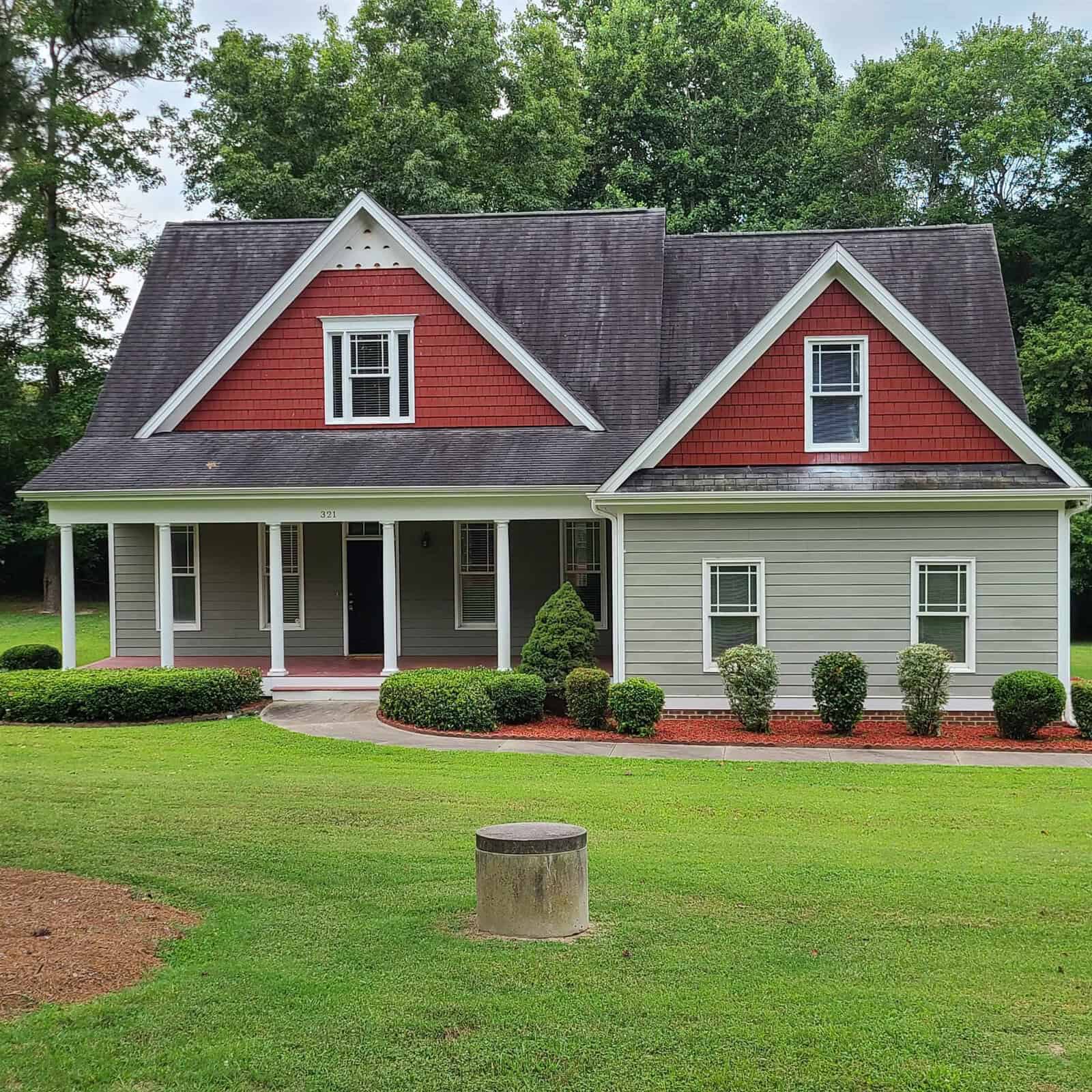 A red and white suburban house at 321 Hedrick Drive, Henderson NC, with a gabled roof, featuring a front porch and manicured bushes, set against a backdrop of green trees and a