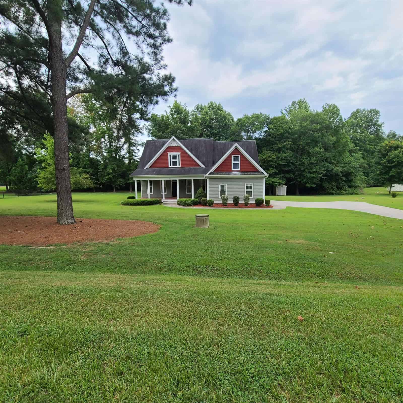 A two-story house with a red roof and white siding, featuring a covered porch with red chairs, surrounded by a well-manicured lawn and trees under a cloudy sky.