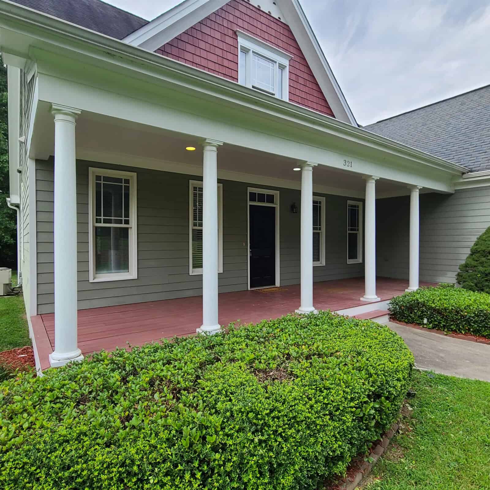 A single-story red house with a covered porch and white columns, featuring a black front door and surrounded by trimmed green shrubbery.
