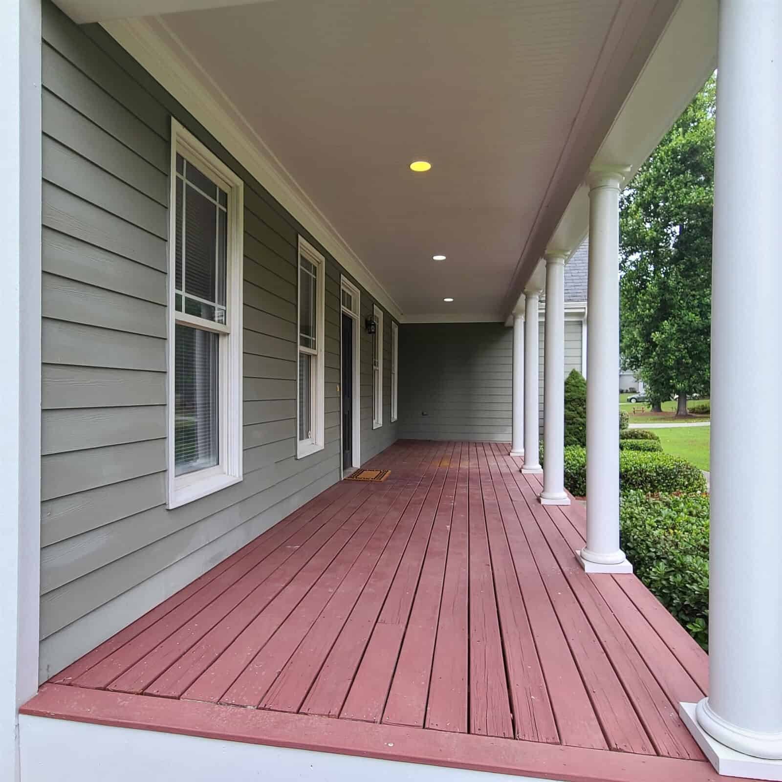 A residential porch featuring red decking, white columns, and gray siding, with a well-maintained lawn visible in the background.
