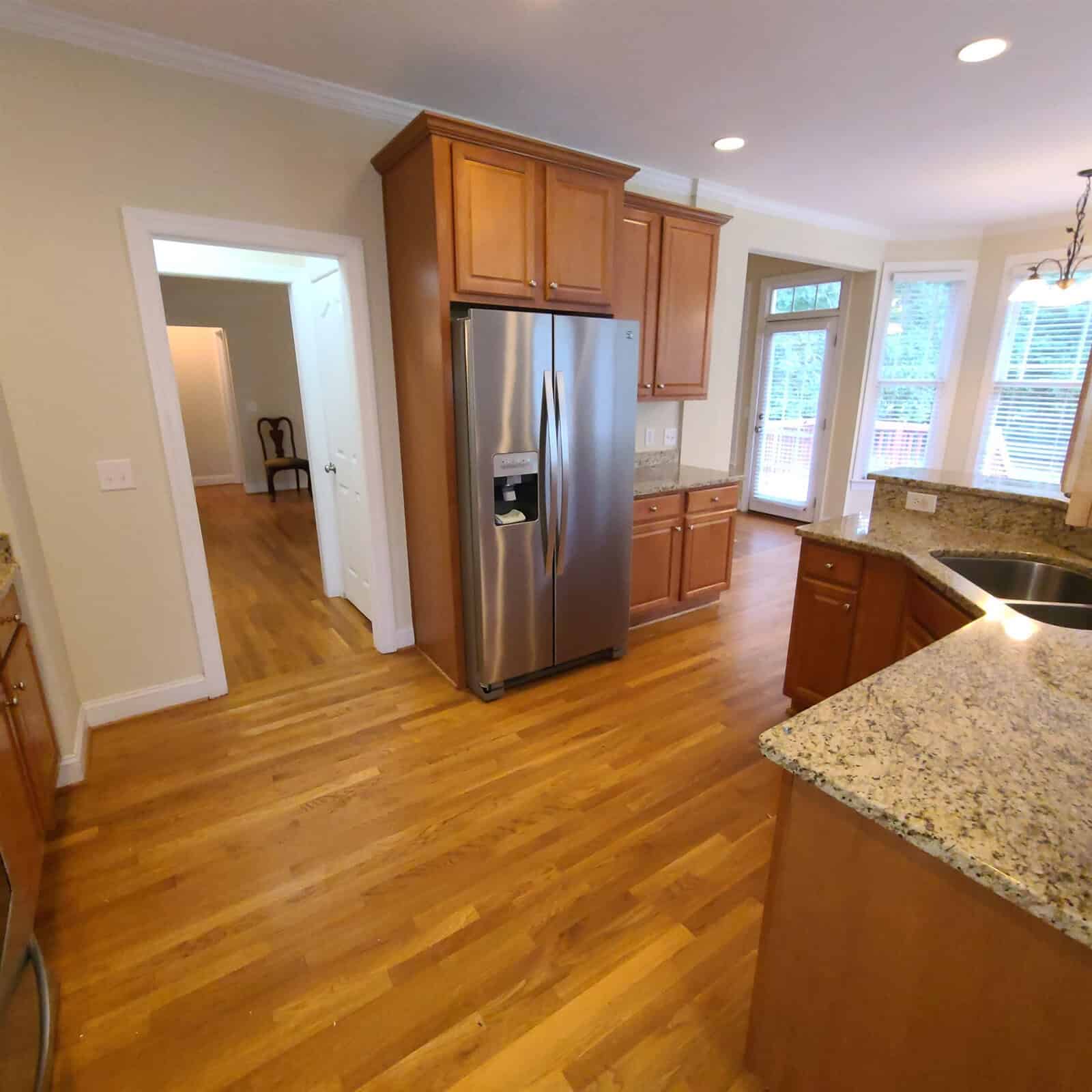 A modern kitchen interior featuring wooden cabinets, stainless steel refrigerator, hardwood floors, and a granite countertop island.