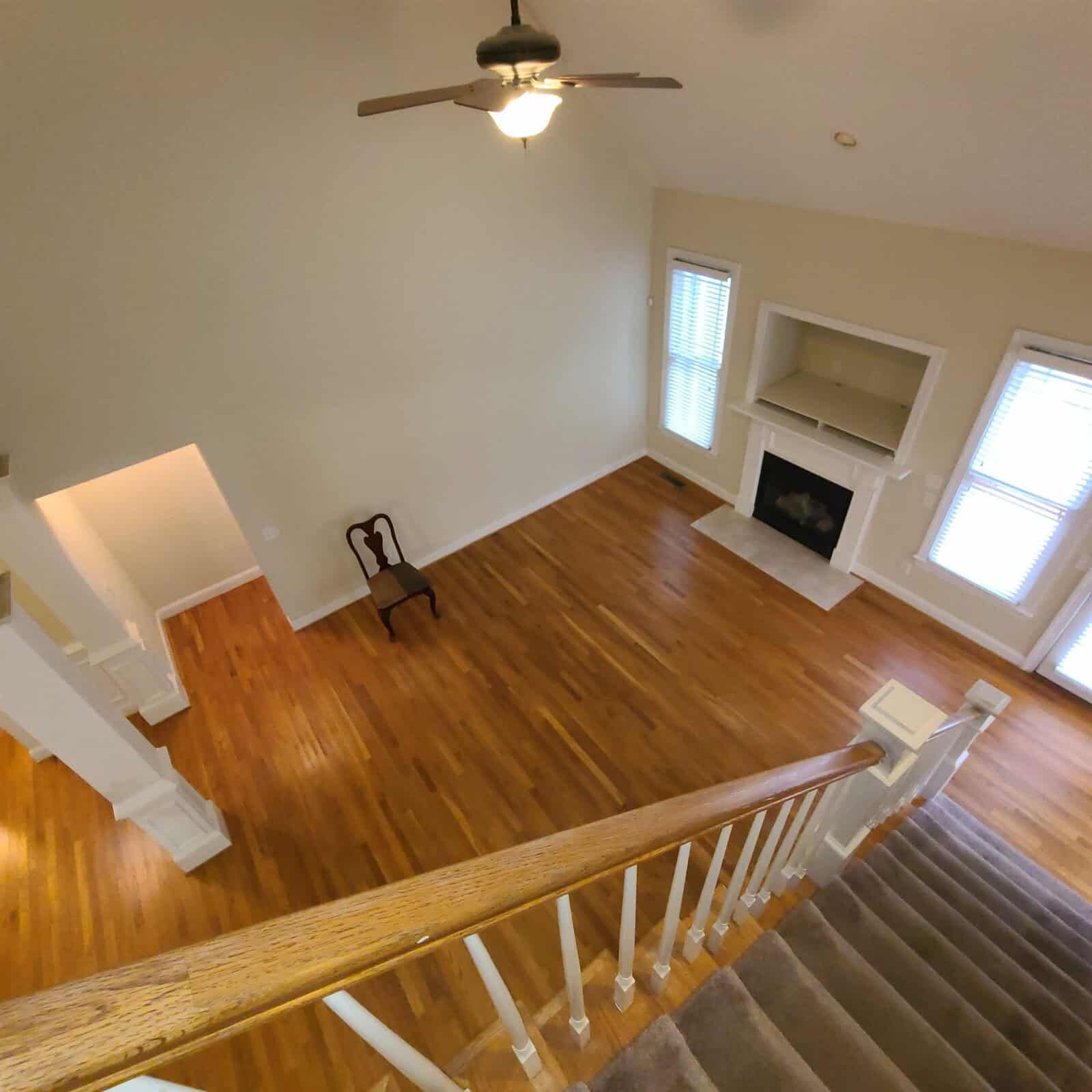 An empty living room with hardwood floors, a fireplace, and a single chair positioned near a wall, viewed from an upper floor landing with white balustrades.