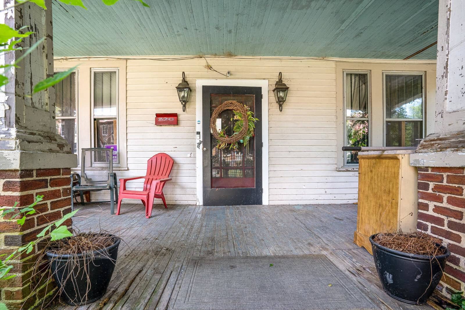 A well-worn front porch with a red chair, wooden door adorned with a wreath, and two potted plants on either side.