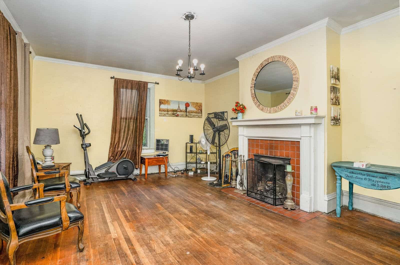 A spacious living room with a hardwood floor, featuring a fireplace, a round mirror above it, a ceiling light fixture, and an assortment of furniture and personal items scattered around the space.
