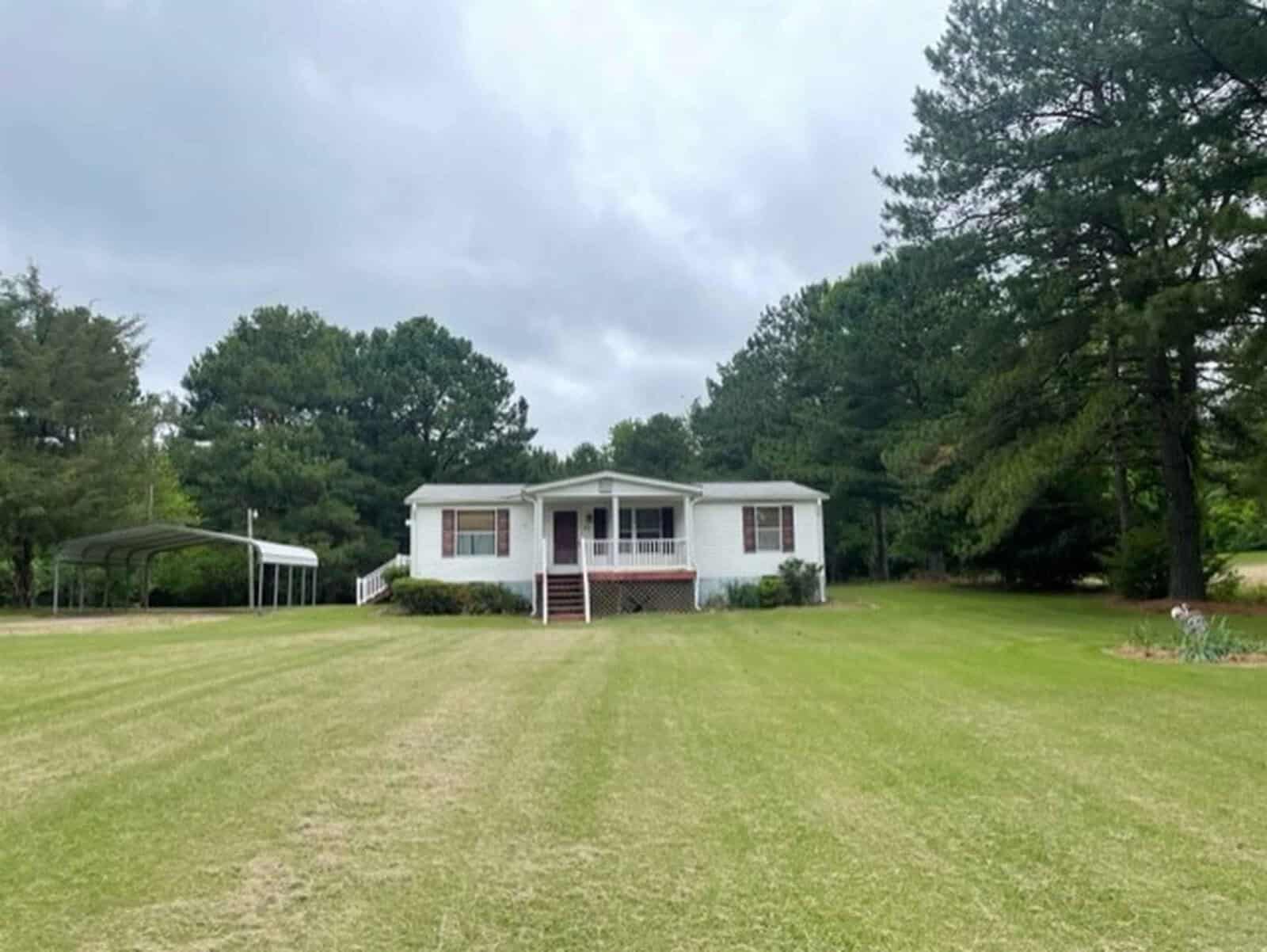 A single-story white house with a red roof and front steps, set against a backdrop of trees under a cloudy sky, with a freshly mowed lawn in the foreground.