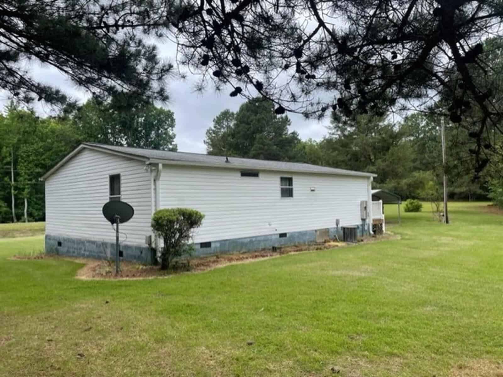 A single-story mobile home with a white exterior and blue trim, situated in a grassy area with trees around, under overcast skies.