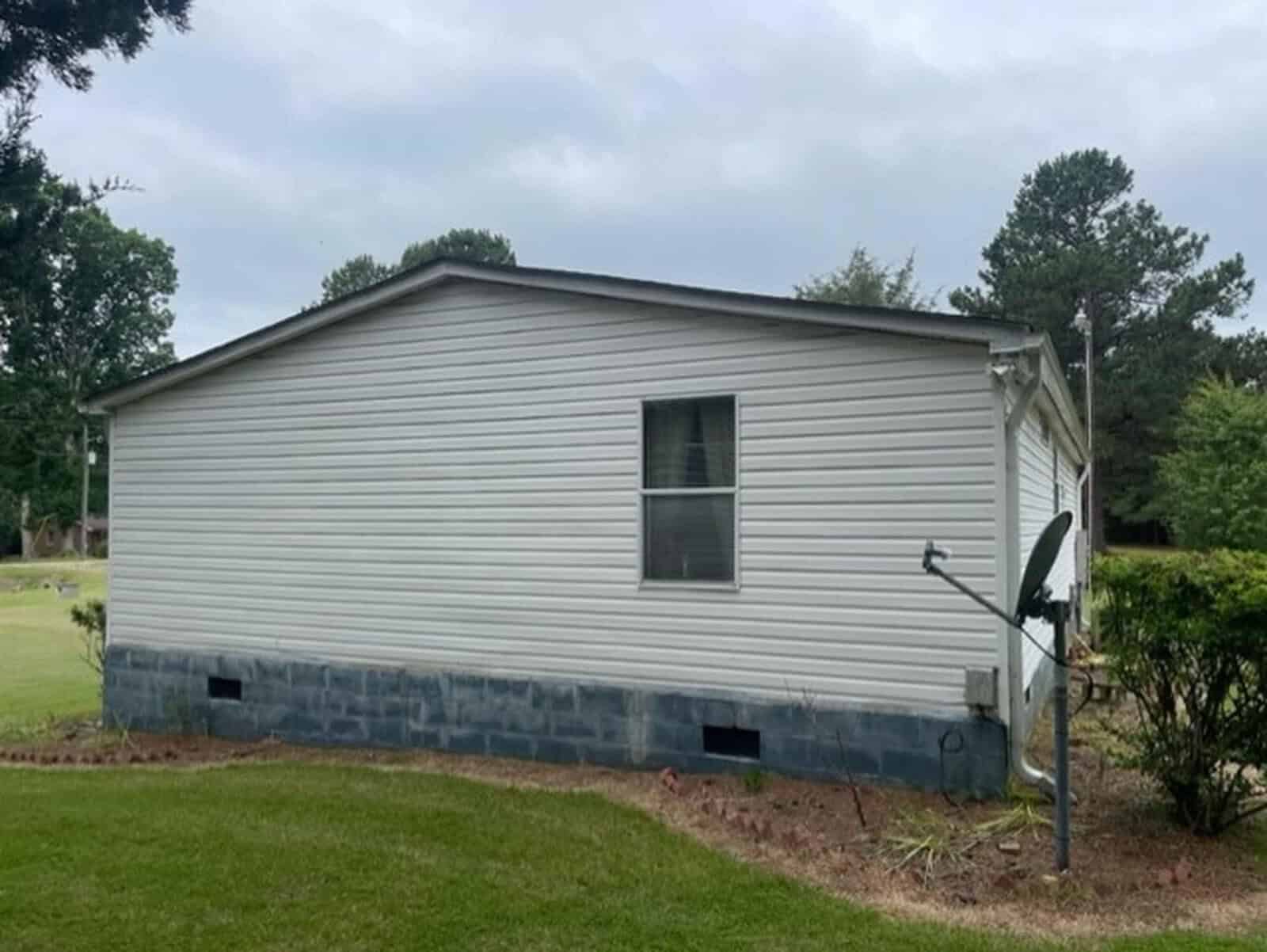 A single-story mobile home with white siding and a dark roof, featuring a side window and a satellite dish mounted in the front yard.