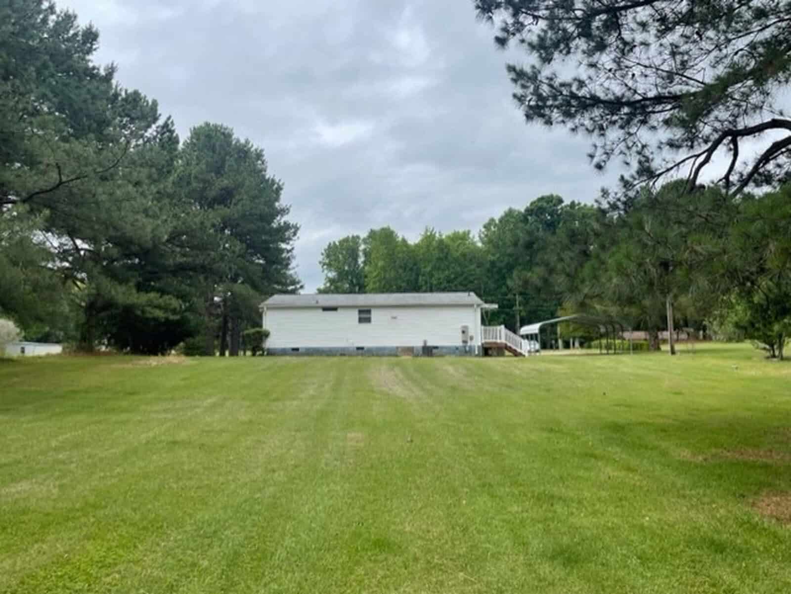 A white single-wide mobile home surrounded by trees with a well-maintained lawn in the foreground.