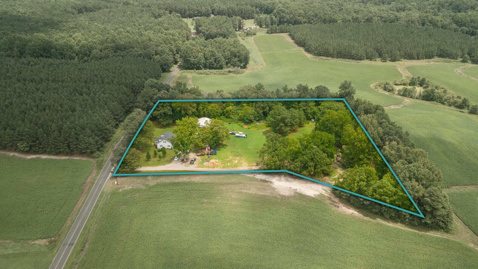 Aerial view of a rural property outlined in blue, showing a cluster of buildings amidst trees, surrounded by open fields and forest.