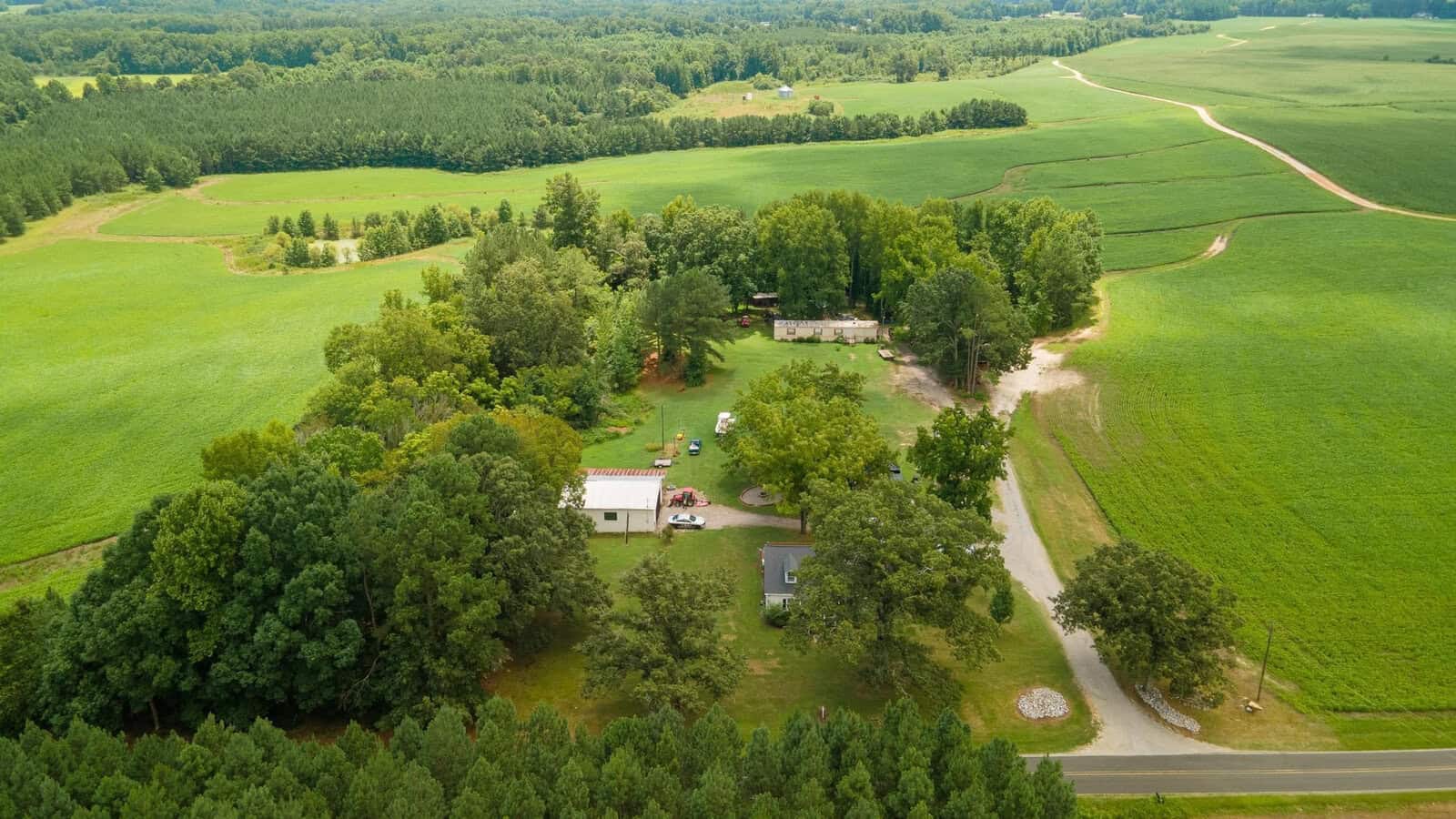 Aerial view of a rural landscape featuring fields, a house with outbuildings, surrounded by trees, and a curved driveway.