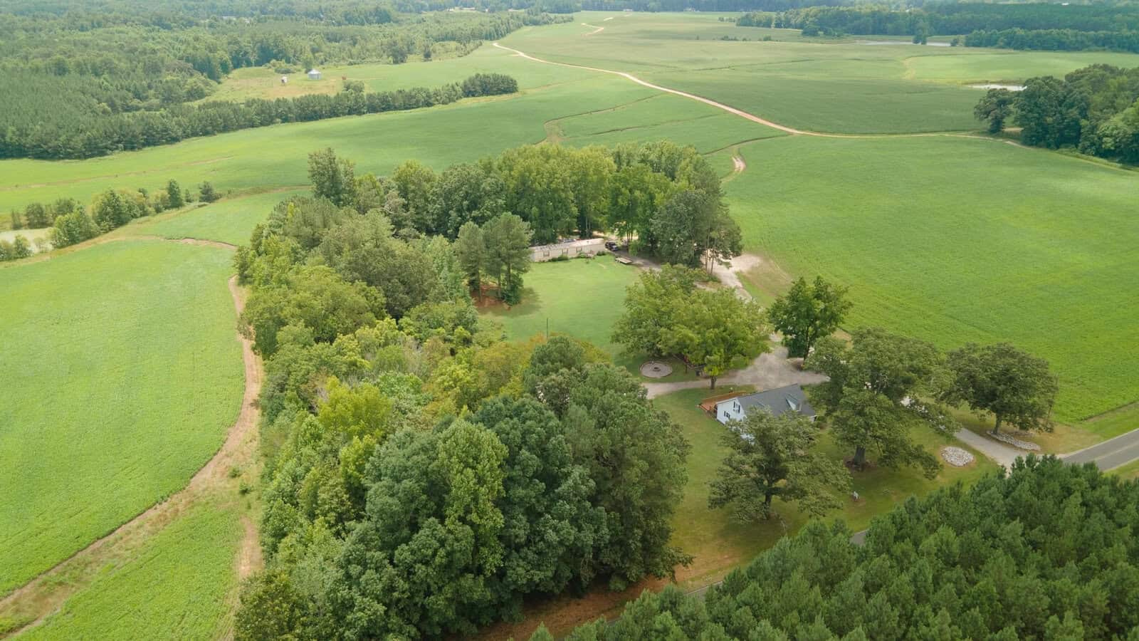 Aerial view of a rural landscape with green fields, a few clusters of trees, and a small residential area with buildings and a driveway.