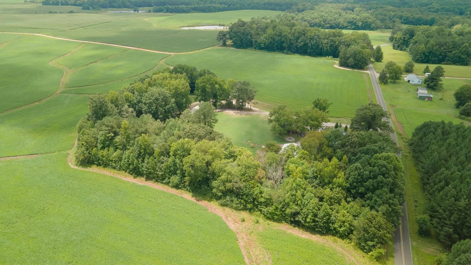 Aerial view of a rural landscape showing fields, a road, and a small cluster of buildings surrounded by trees.
