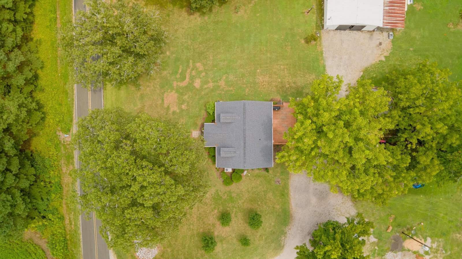 Aerial view of a single-family house with a shingle roof, surrounded by green trees and a lawn, with a dirt driveway on the right and a roadway on the left.