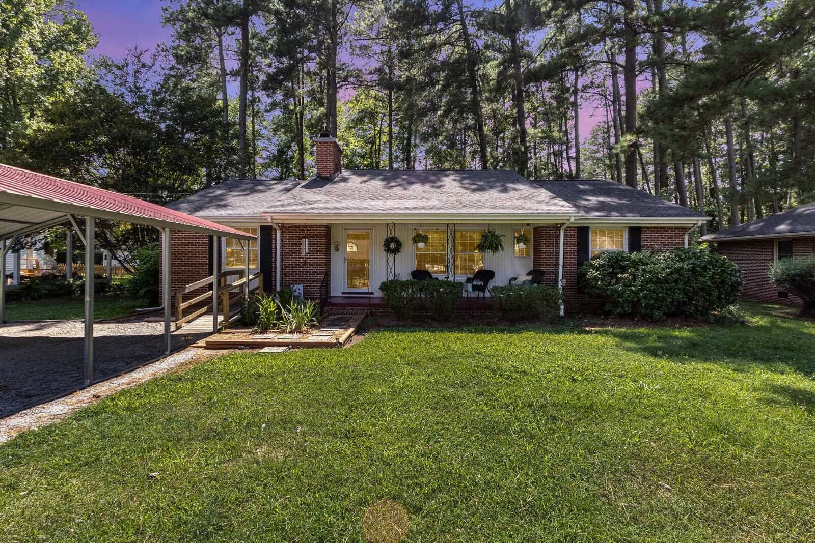 A single-story suburban house with a brick facade, white trim, and a covered front porch, surrounded by tall pine trees and a well-maintained lawn.