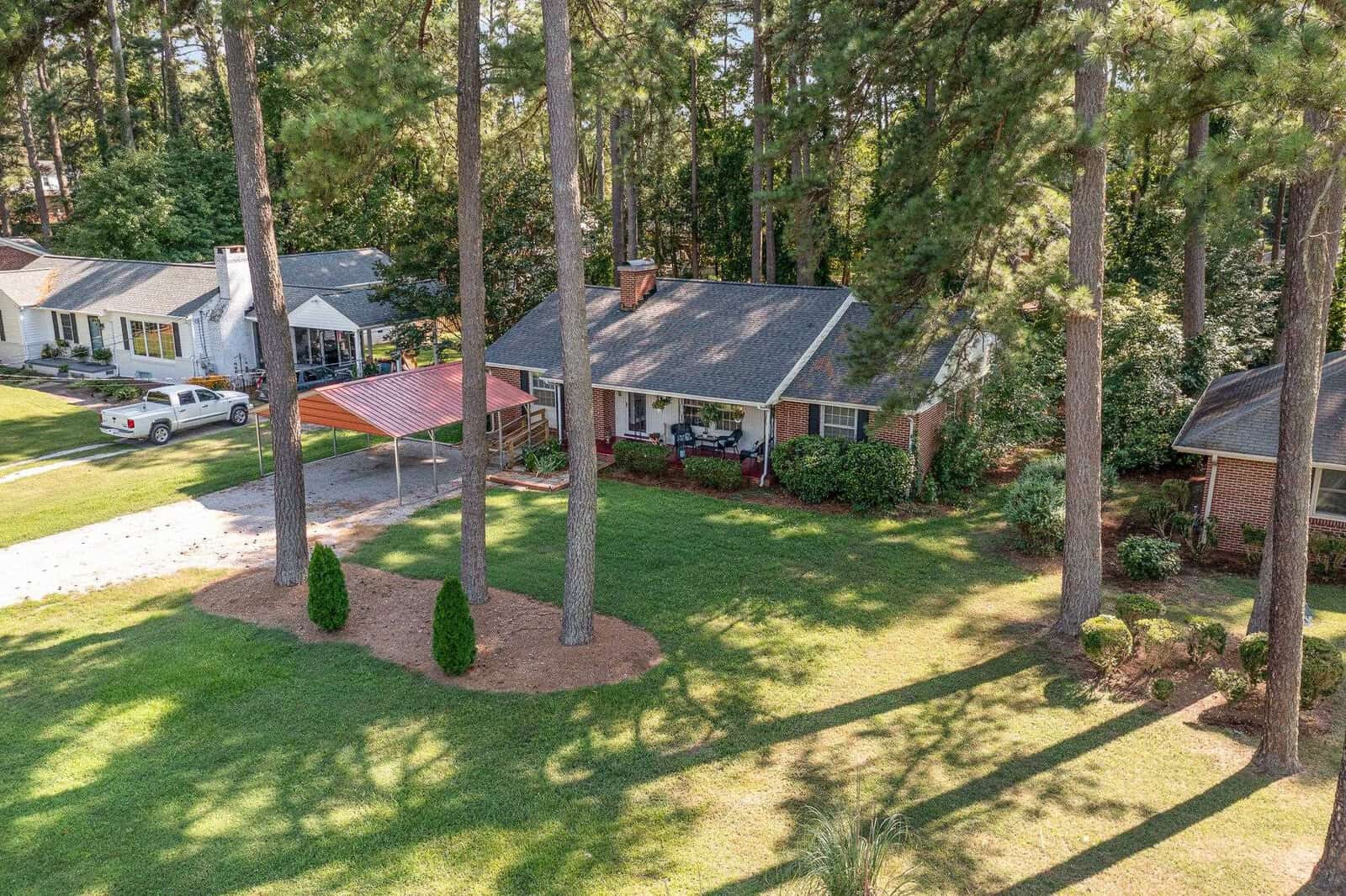 Aerial view of a residential neighborhood showing a single-story house with a brown roof, a car parked in the driveway, surrounded by tall pine trees and well-manicured lawns.