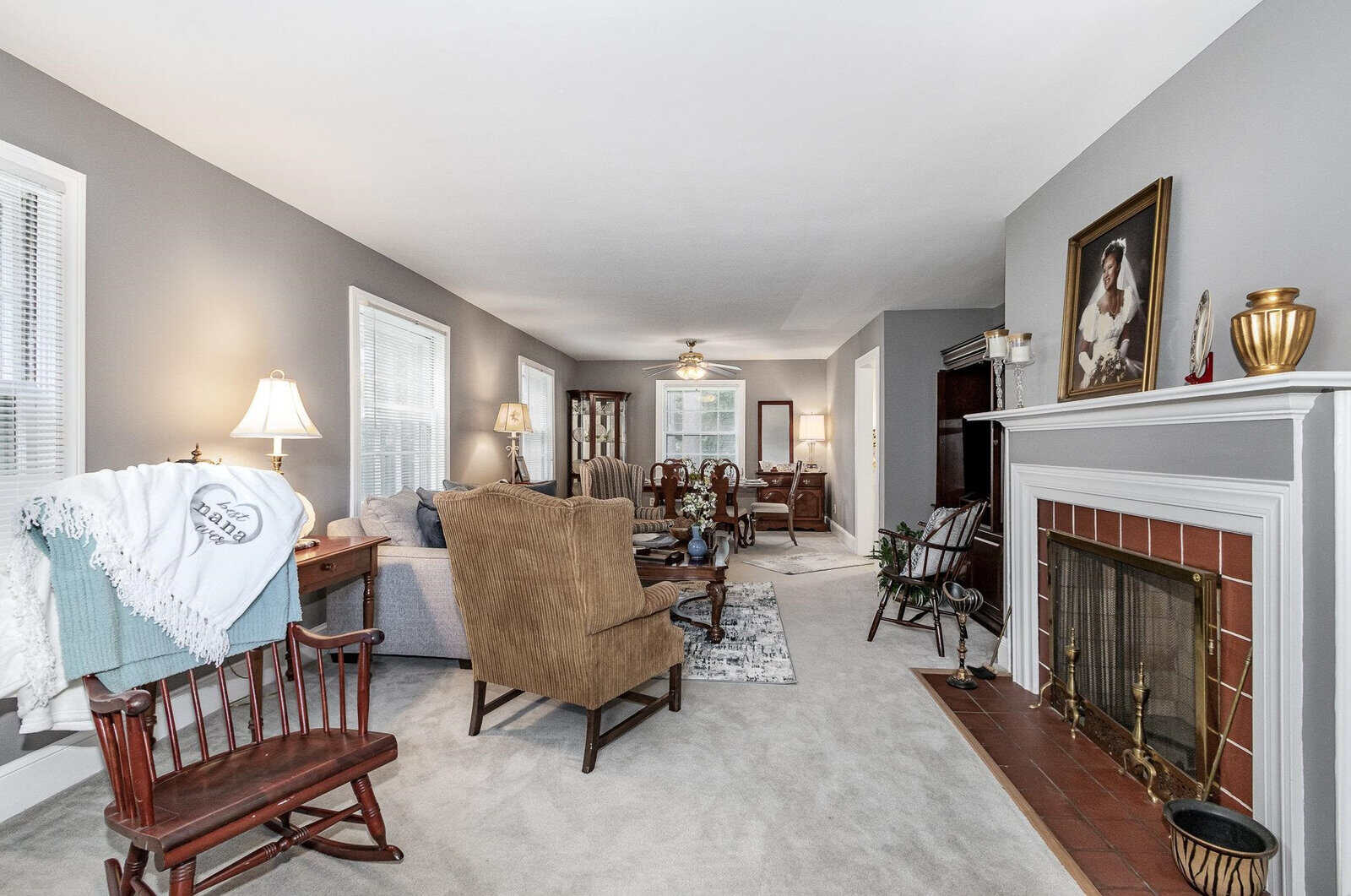 A well-lit, spacious living room with a fireplace, rocking chair, armchair, and decorative items, featuring neutral colors and a portrait above the mantel.