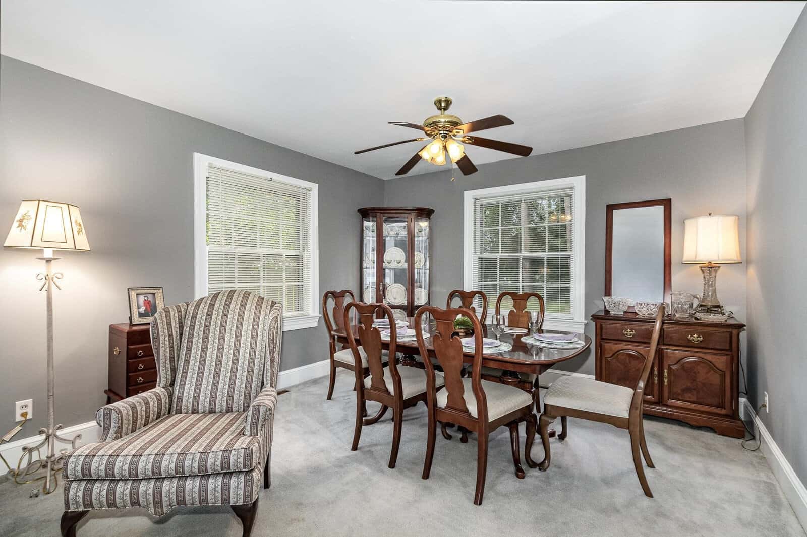 A well-lit, traditional dining room featuring a wooden table with chairs, an upholstered armchair, a sideboard, two floor lamps, and a ceiling fan, with natural light coming in through two windows and a glass door.